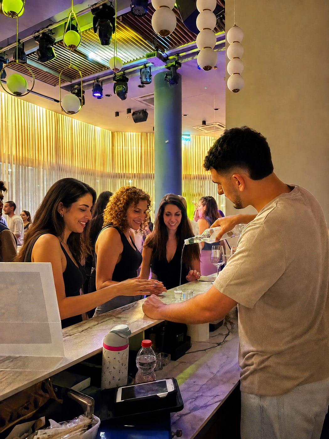 ברמן מוזג לשלוש נשים בבר השיש, מתחת לתאורת מסיבה צבעונית